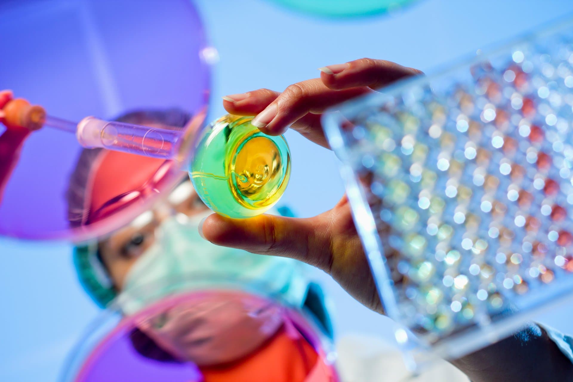 Researcher in mask and cap examining a green and yellow liquid sample in a flask alongside a multi-well testing tray in a lab.